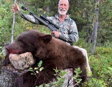 Hunter smiling with a rifle and a harvested color-phase black bear in a lush Wyoming forest.