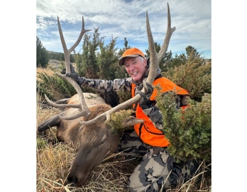 A beaming hunter in a camouflage jacket and orange vest, holding the antlers of a large bull elk harvested on a guided SNS hunt in Wyoming.