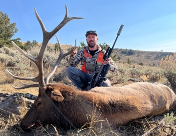 A hunter seated next to a harvested bull elk, with a rifle resting nearby. The elk's antlers extend majestically against the backdrop of Wyoming’s scenic wilderness.