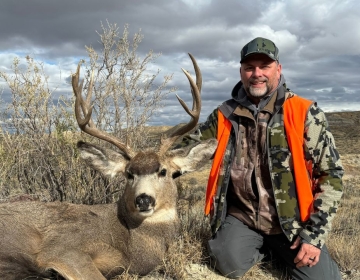 Hunter in an orange vest and camo jacket kneeling beside a harvested mule deer with impressive antlers under cloudy Montana skies.