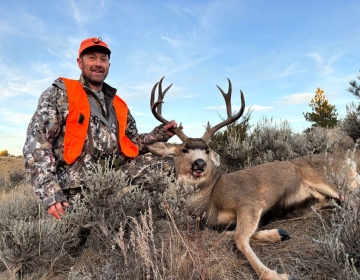 Hunter in an orange vest sitting beside a harvested mule deer in a sagebrush-covered Montana landscape with a blue sky.