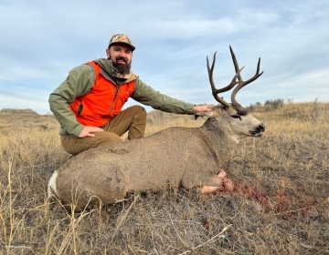 A hunter wearing a bright orange vest and camouflage jacket kneeling beside a mule deer buck in an open Wyoming field.