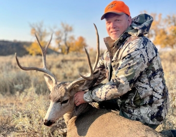 A hunter shows off a mule deer buck with impressive antlers, harvested during a hunt with SNS Outfitter & Guides amidst Wyoming’s fall foliage.