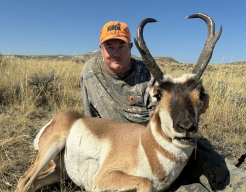 A hunter posing with a pronghorn antelope taken in Wyoming. The antelope has distinct curved horns, and the hunter wears a camouflage outfit and an orange NRA hat, kneeling in the golden grasslands.