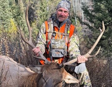 Hunter wearing camouflage and an orange vest holding the antlers of a harvested bull elk in a forested area during a Wyoming elk hunt.