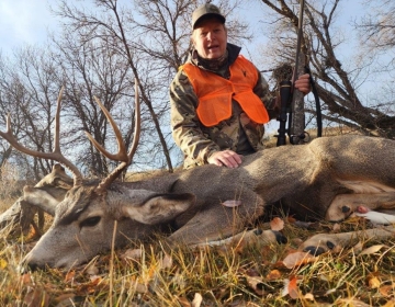 A hunter in a camo jacket and orange vest sitting beside a large mule deer buck in a golden autumn landscape.