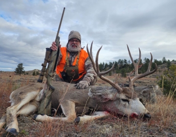 Hunter in an orange vest holding a rifle and posing beside a harvested mule deer with large antlers in a rugged Montana field.