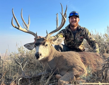 A young hunter in camouflage gear and sunglasses kneeling behind a mule deer with a striking rack, framed by Wyoming’s rugged terrain and clear skies.