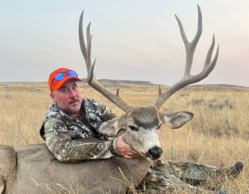 A satisfied hunter displays a large mule deer buck taken during an SNS-guided hunt in Wyoming's golden grasslands, framed by the expansive horizon.