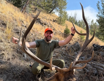 Hunter in an orange hat posing beside a large bull elk on a steep hillside during a sunny Wyoming elk hunt.