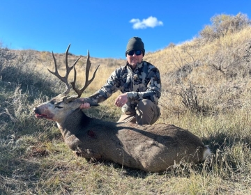 Hunter in camouflage posing with a harvested mule deer on a grassy Montana hill under a bright blue sky.