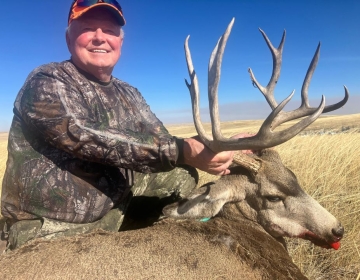 Hunter wearing camouflage holding the antlers of a large mule deer buck with a backdrop of Wyoming's open plains.