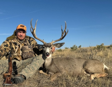 A hunter with a mule deer harvested in Wyoming, sitting proudly beside the animal in a dry, open field. The deer features a symmetrical set of antlers, and the hunter is dressed in camouflage with an orange NRA hat, holding a wooden-stock rifle.