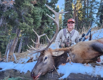 Smiling female hunter in winter gear posing with a harvested bull elk in a snowy forest during a successful Wyoming rifle hunt.