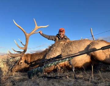 A hunter in an orange hat posing with a massive bull elk lying in the Wyoming grasslands alongside his rifle, showcasing a successful hunt.