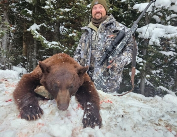 Hunter in camouflage standing in snow next to a harvested black bear, highlighting an early springtime hunt in a snowy Wyoming forest.