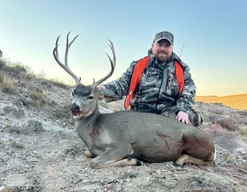 Hunter in orange and camouflage next to a mule deer buck on rocky Wyoming terrain, highlighting a guided hunt success.