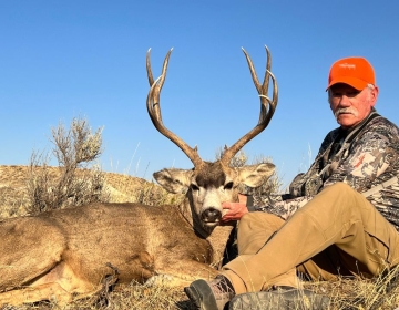 A proud male hunter wearing an orange hat displays his mule deer, harvested during a guided hunt with SNS Outfitter & Guides under clear Wyoming skies.