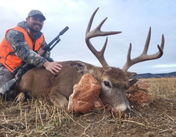 Hunter in an orange vest holding a rifle posing with his 8 point whitetail deer