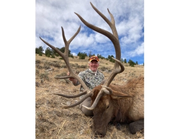 A hunter smiling with a 6x6 bull elk, posing on a hillside covered with dry grass under a partly cloudy sky.