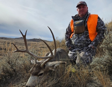 Hunter in camouflage with bright orange safety gear kneeling beside a mule deer harvested during a guided hunt in Wyoming's rugged terrain.