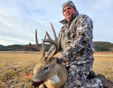 Hunter posing with a freshly harvested whitetail deer in a wide open field, showcasing a successful two-state hunt experience.