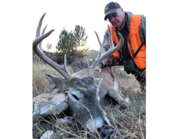 Hunter in an orange vest and camo gear leaning over a harvested whitetail deer, with the sun setting behind Wyoming's rugged terrain.