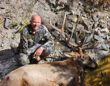 Hunter in a rocky area posing with a large bull elk on the ground, marking a successful guided elk hunt in Wyoming.