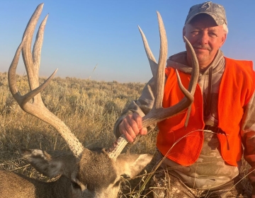 Hunter kneeling beside a trophy mule deer buck with tall antlers in the open plains, showcasing a successful Wyoming hunt.