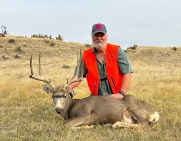 Hunter wearing an orange safety vest posing with a large mule deer buck harvested during a guided hunt in a grassy Wyoming field.