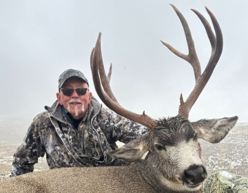 Hunter wearing camouflage jacket and smiling with his mule deer after the hunt in a snowy Wyoming landscape