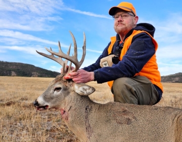 A hunter in an orange cap and vest posing with a mature whitetail deer against a backdrop of expansive Montana plains. Experience the thrill with SNS Outfitter & Guides.