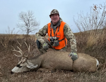 A hunter in a camo outfit and orange vest smiling beside a whitetail deer buck in a grassy Wyoming setting.