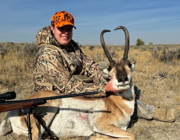 A young hunter wearing camo clothing and an orange hat  proudly poses with a pronghorn antelope in the prairie of Wyoming.