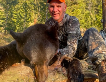 Hunter in orange cap and camouflage gear posing beside a harvested black bear in a sunny Wyoming forest clearing during a successful hunt.