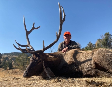 A smiling hunter with an orange hat kneeling behind a massive bull elk, showcasing the animal's impressive antlers on a sunny day in Wyoming.