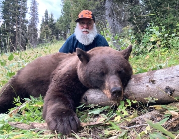 Hunter with a long white beard posing behind a harvested black bear in a lush green forest during a guided hunt.