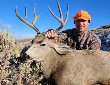 A hunter wearing an orange hat kneeling beside a freshly harvested mule deer buck in the frosty Wyoming landscape.