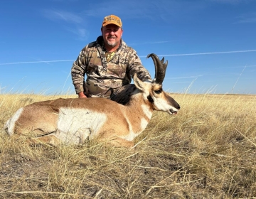 Hunter smiling next to a harvested pronghorn antelope in an open field under a clear blue Wyoming sky.