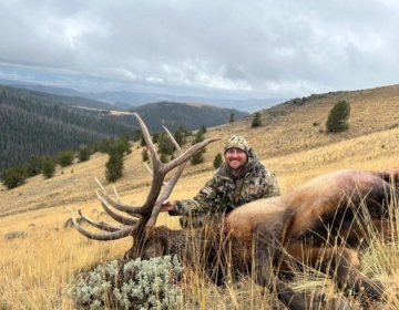 Smiling hunter in camouflage posing with a heavy horned bull elk on a ridge overlooking Wyoming's scenic mountains and valleys.