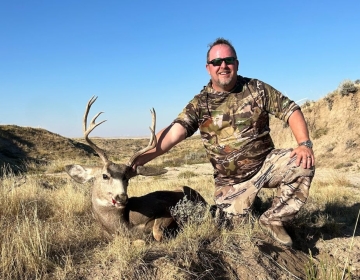 Hunter in camouflage posing with a trophy mule deer buck in rugged Wyoming terrain during a successful hunting trip.