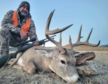 Hunter in camouflage and an orange vest posing with a harvested whitetail deer, rifle resting beside the trophy in a Wyoming field at sunset.